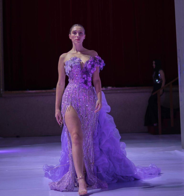 Modelo con vestido lila brillante y corte lateral, caminando en pasarela con expresión de confianza y postura elegante.