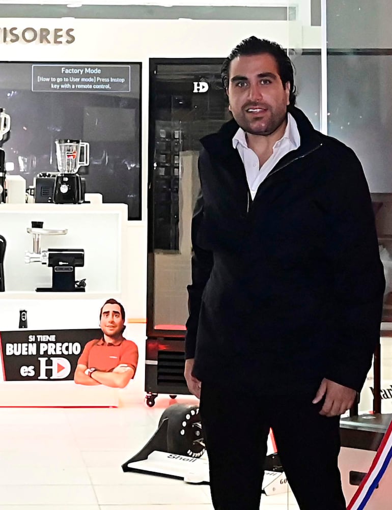 Hamza Damani viste chaqueta negra en una tienda moderna de electrodomésticos, rodeado de licuadoras y carteles promocionales.