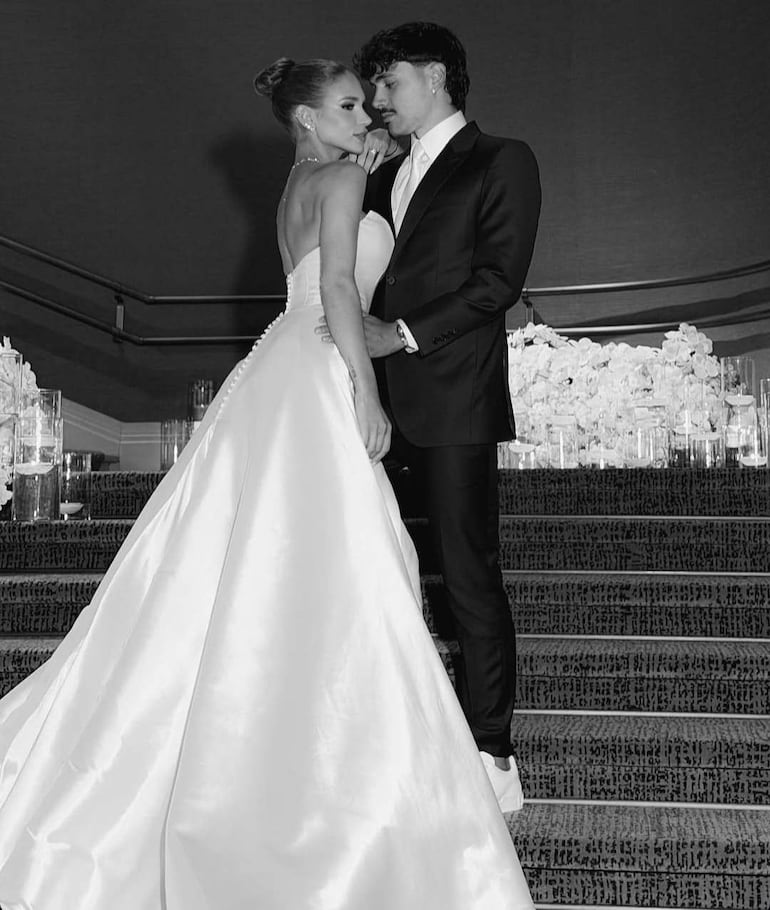 Novia con vestido blanco y cola, mirando al novio vestido con traje negro en una escalera decorada. Ambiente íntimo y elegante.