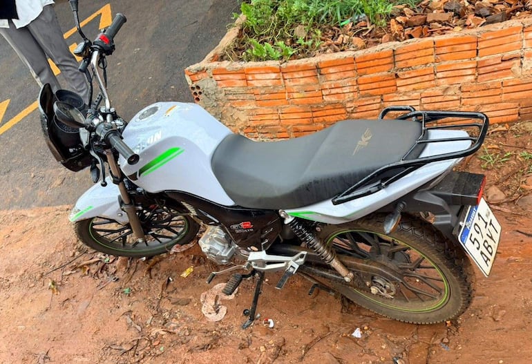 Motocicleta gris con detalles en verde aparcada en terreno arenoso, rodeada de hojas secas y un muro de ladrillos.