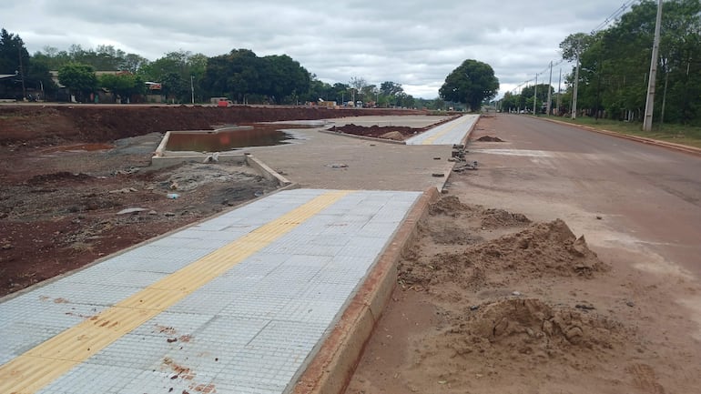 Obra en construcción en ruta, senda peatonal parcialmente terminada y cuerpo de agua visible en un día nublado.