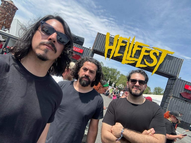 Tres hombres con camisetas negras y gafas de sol en el festival Hellfest, sonriendo en un ambiente festivo.