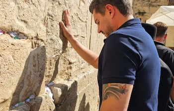 Hombre de perfil con camiseta oscura y tatuaje visible, tocando la Muralla de las Lamentaciones con expresión concentrada.