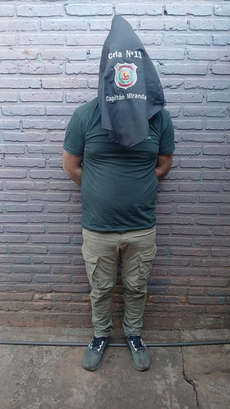 Hombre de espaldas con capucha negra y bordados 'Cría No 11' y 'Capitán Miranda', en ambiente de detención con pared de ladrillo.