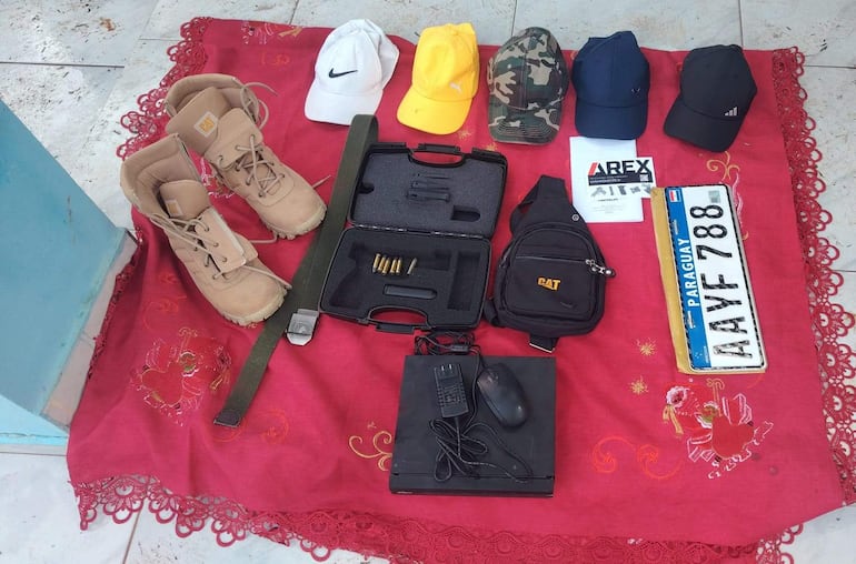 Objetos incautados sobre manta roja: gorras de varios colores, botas beige, un estuche negro y una matrícula paraguaya.