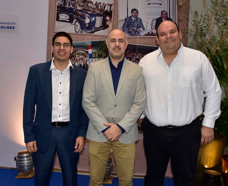Tres hombres sonrientes en trajes formales, posando en un salón elegante decorado para el aniversario de La Consolidada.