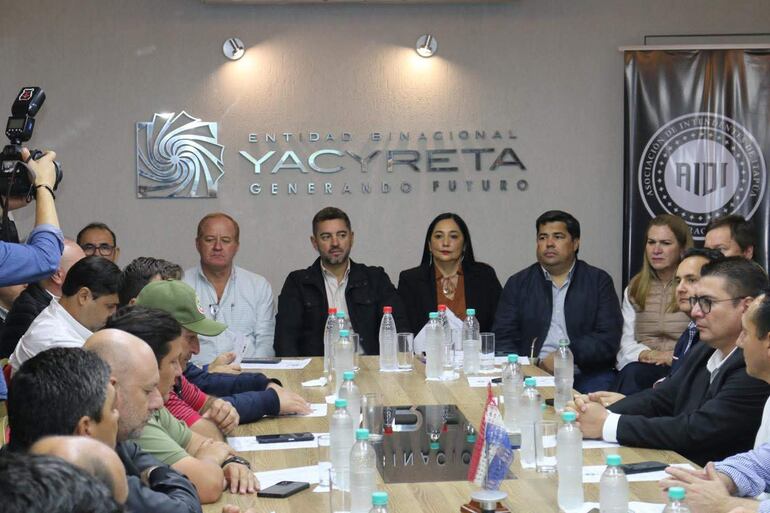 Alliana, en traje oscuro, escucha atentamente a los intendentes, Pereira Rieve y Barán, en ambiente formal con botellas de agua y papeles sobre la mesa. Reunión en la Entidad Binacional Yacyretá.