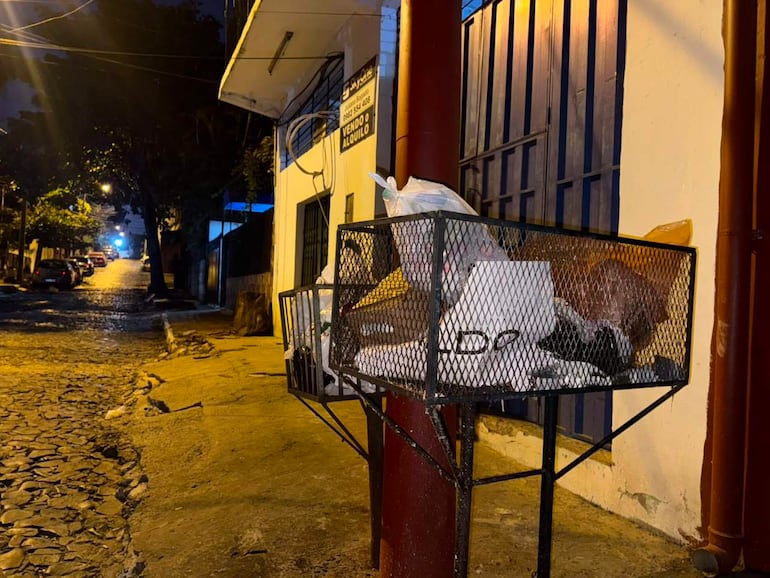 Canasto de metal lleno de bolsas de basura en calle empedrada, sin personas visibles, iluminado por luces de edificios.