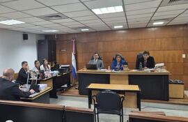 Sala de audiencias con grupo de abogados, una mujer con blusa azul hablando y un hombre de saco negro, todos en actitud formal.