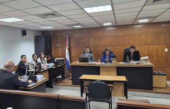 Sala de audiencias con grupo de abogados, una mujer con blusa azul hablando y un hombre de saco negro, todos en actitud formal.