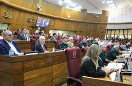 Personas en trajes formales escuchan atentamente al orador en un recinto legislativo con pantalla.