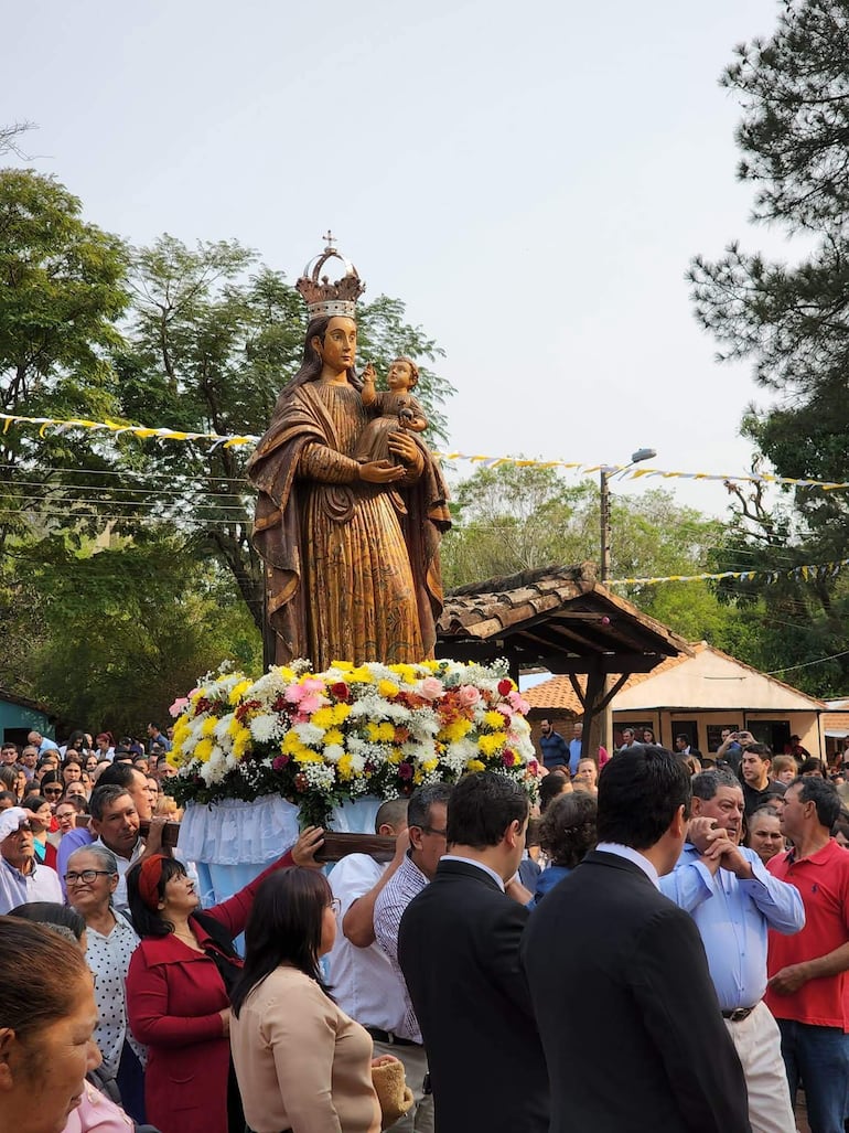 Concurrida celebración al aire libre con imagen de la Virgen María adornada con flores, rodeada de fieles con expresiones de devoción.