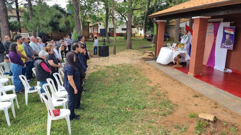 Grupo de personas reunidas frente a un altar decorado con flores y retratos, mientras un sacerdote sostiene la bandera de Paraguay.