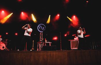 Mau y Ricky en el escenario, ambos con camisetas blancas, cantando en medio de luces rojas y amarillas mientras la multitud los aplaude.