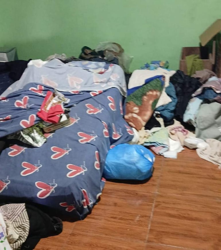 Ambiente interior desordenado con camas, mantas sobre el suelo y objetos revueltos, sin personas visibles.