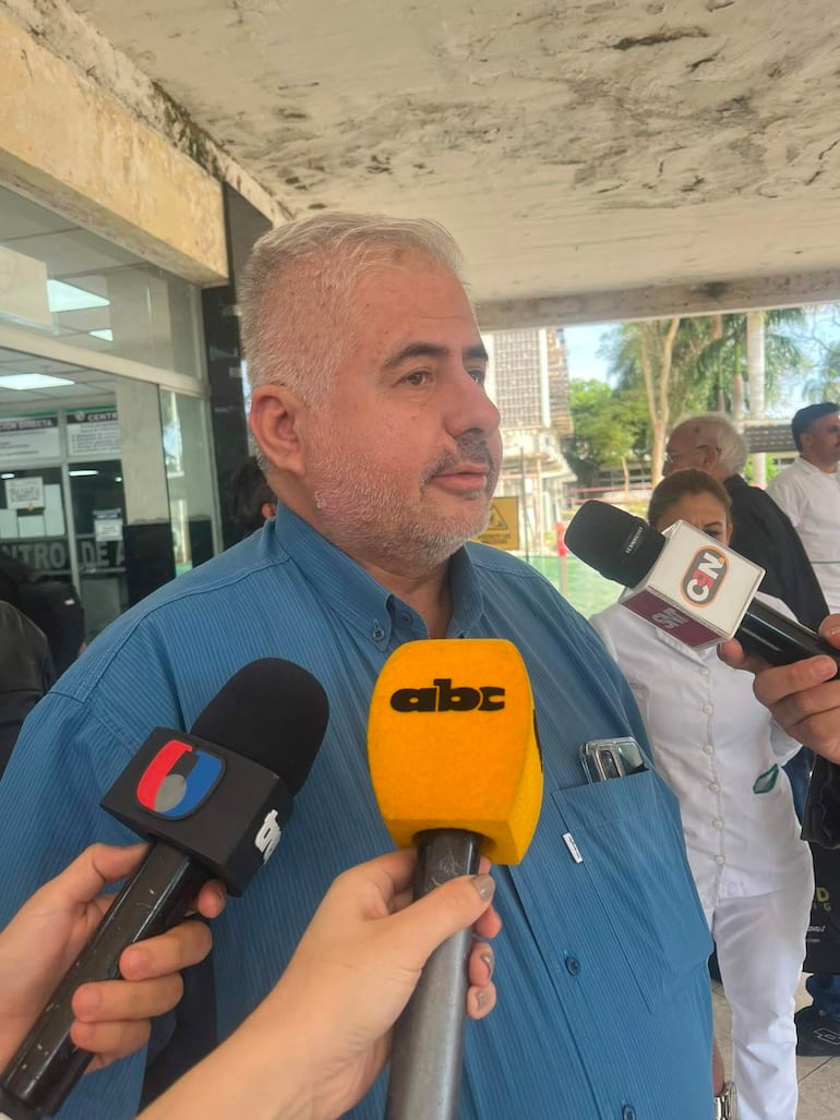 Hombre de cabello gris y barba, en camisa azul, rodeado de micrófonos en un evento al aire libre, con edificios y árboles al fondo.