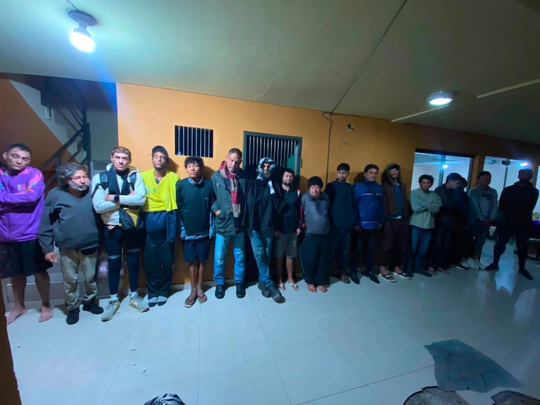 Grupo de 15 hombres alineados en sala con paredes naranjas, algunos conversando y otros con actitud seria.
