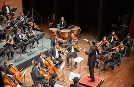 Conjunto orquestal de 20 músicos en trajes formales negros, director al centro usando batuta en un auditorio iluminado.