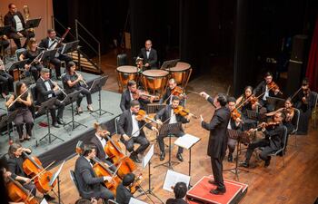 Conjunto orquestal de 20 músicos en trajes formales negros, director al centro usando batuta en un auditorio iluminado.