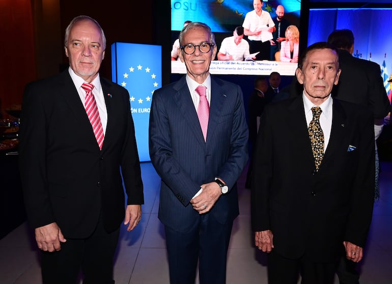 Tres hombres de pie en evento formal, uno con chaqueta azul y corbata rosa, en ambiente decorado con pantalla de fondo.
