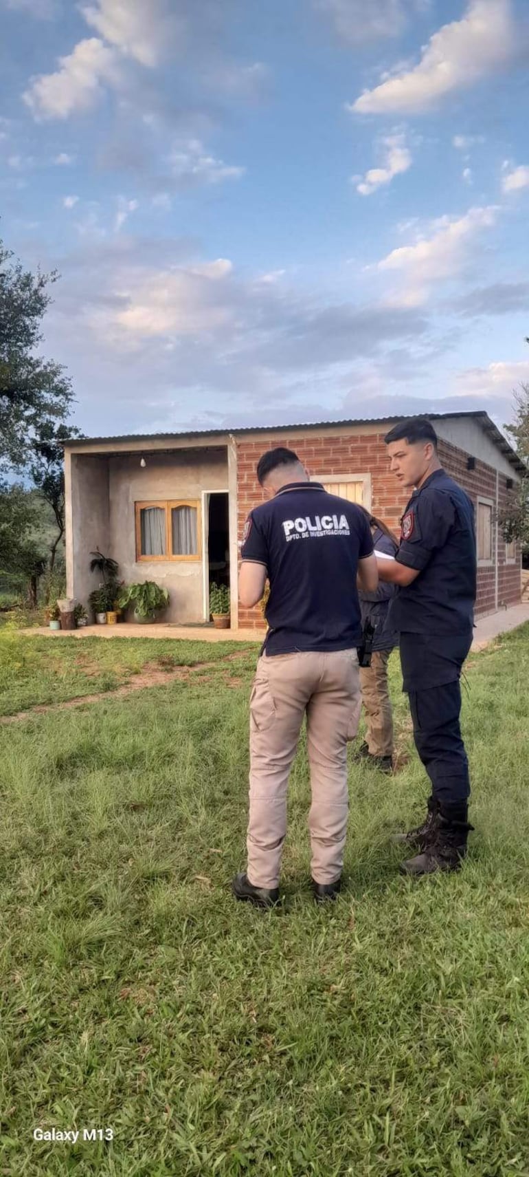 Dos policías conversan frente a una casa de ladrillos, uno toma notas mientras el otro observa, en un ambiente rural con césped.