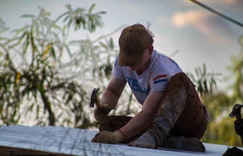 Hombre con gorro marrón y camiseta blanca trabaja concentrado con un martillo en un techo rodeado de vegetación.