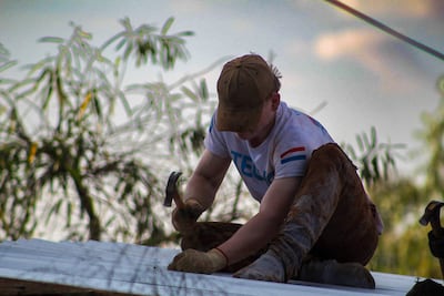 Hombre con gorro marrón y camiseta blanca trabaja concentrado con un martillo en un techo rodeado de vegetación.