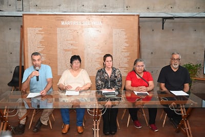Hombre en camiseta azul habla con micrófono en mesa con documentos, rodeado de mujeres con ropa clara y oscura en ambiente serio.