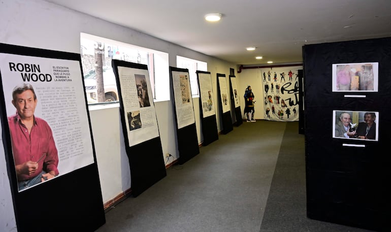 Cartel con imagen de Robin Wood y paneles informativos, con público observando en ambiente interno iluminado.