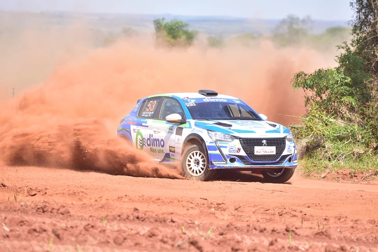 Peugeot de rally en movimiento, con colores azul y blanco, levantando polvo en un camino rural con vegetación al costado.