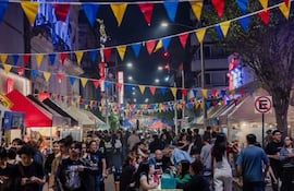 Gente celebrando en la calle iluminada, con trajes informales y puestos de comida decorados con banderines rojo, azul y amarillo.