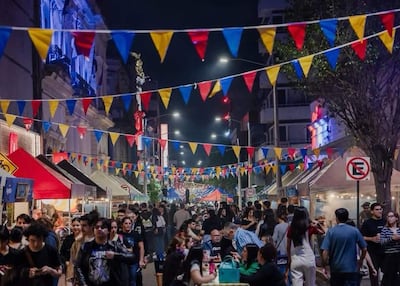 Gente celebrando en la calle iluminada, con trajes informales y puestos de comida decorados con banderines rojo, azul y amarillo.