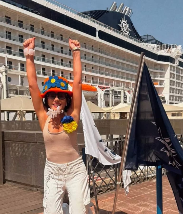 Mujer con sombrero colorido y camiseta sin mangas sonríe, levantando los brazos frente a un gran barco de crucero.