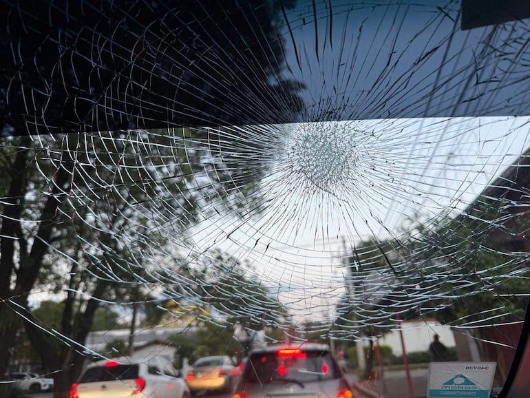 Parabrisas fracturado con grietas visibles, vista desde el interior del vehículo hacia automóviles detenidos en la calle.