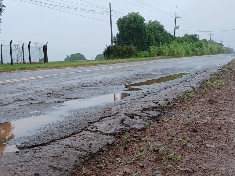 Carretera rural con pavimento desgastado y charcos de agua, rodeada de vegetación y cerco de alambre en un día de lluvia.