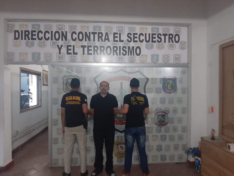 Tres hombres en oficina de seguridad, dos de espaldas con camisetas negras de Policía Nacional y uno al centro con gafas y barba.