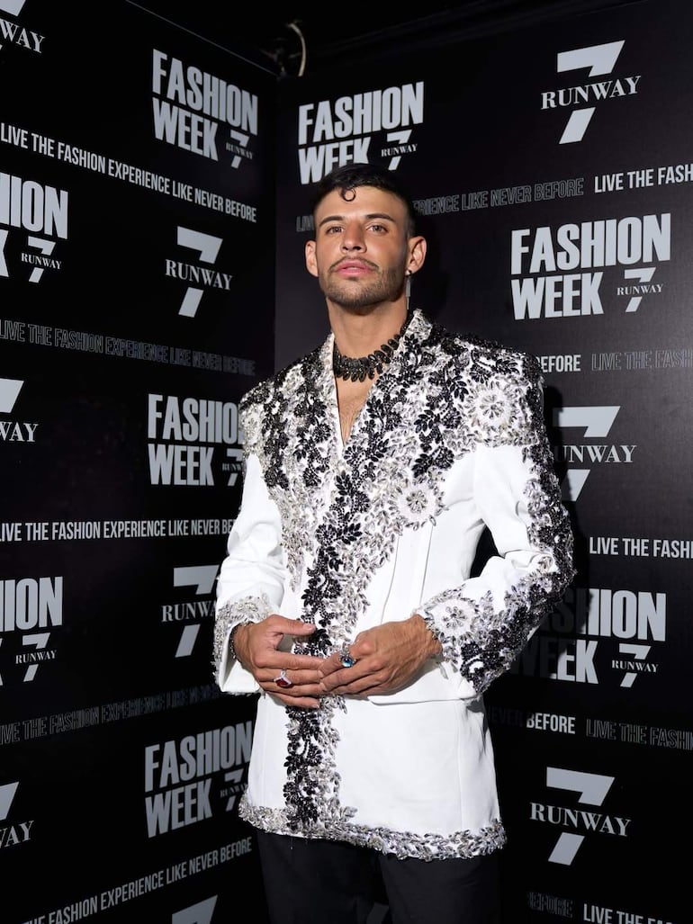 Un hombre de pie en primer plano, con abrigo blanco y detalles en negro y plata, posando en un evento de moda.