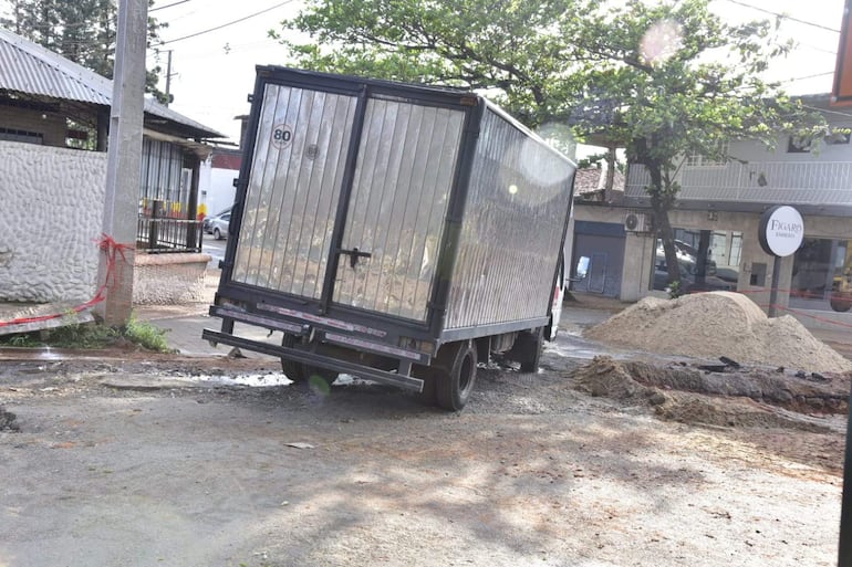 Camión metálico estacionado en calle con construcción, entorno con montículo de arena y cintas de advertencia visibles.