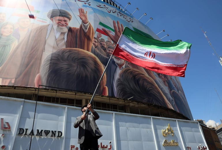 Un iraní porta una bandera de su país frente al mural con la imagen del fallecido ayatolá Alí Jamenei bajo fuego iraní.
