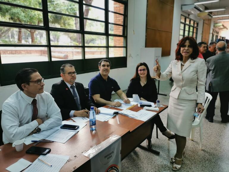 Mujer de pie con traje claro sostiene una tarjeta, rodeada de hombres sentados en un ambiente iluminado y de trabajo.