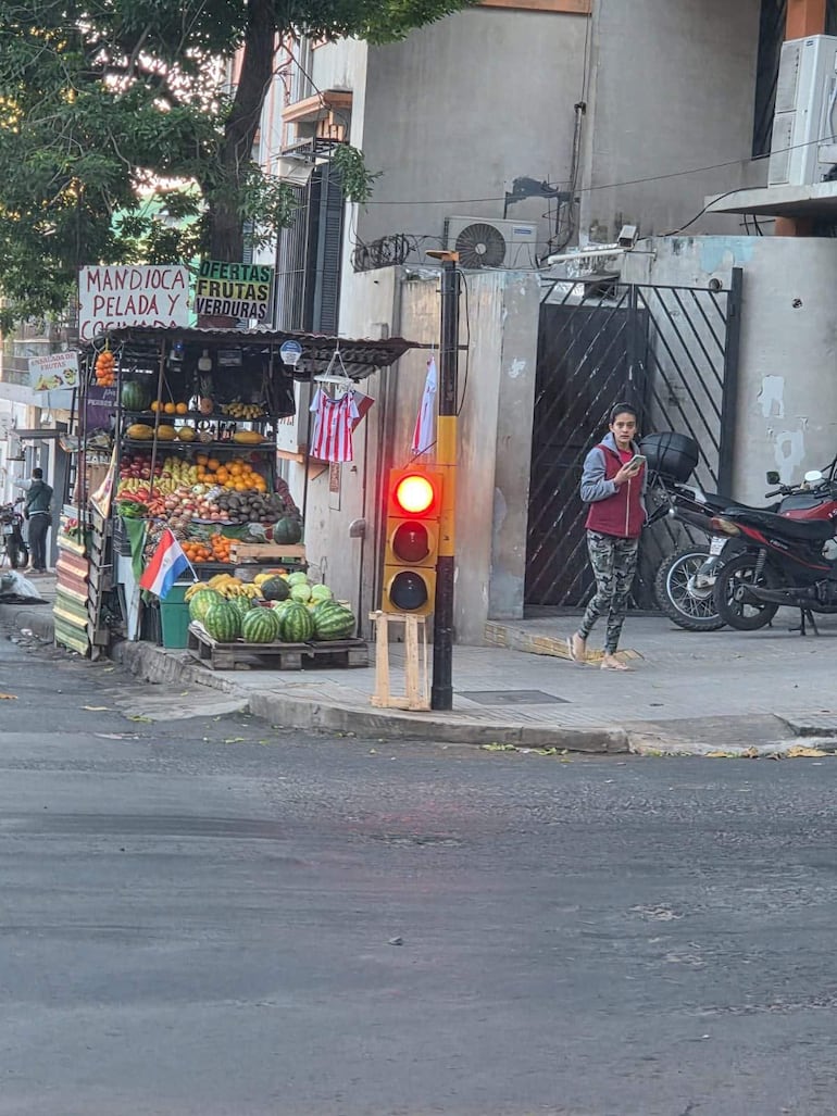 Mujer con suéter gris y pantalones camuflados usa su teléfono frente a un semáforo y un puesto de frutas.