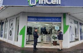 Policía en uniforme frente a la tienda InCell, clima lluvioso con publicidad de productos tecnológicos visible.