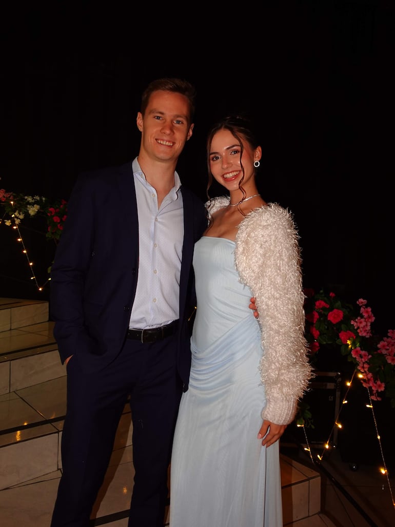 Joshua Duerksen en traje oscuro sonríe junto a Anneke Rahn con vestido azul claro y chaqueta blanca, en ambiente festivo.
