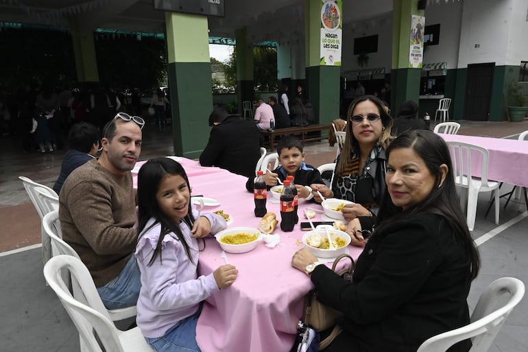 Grupo de cuatro personas sonrientes alrededor de mesa con mantel rosa, comiendo y conversando en un comedor escolar.