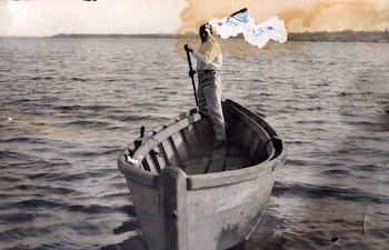 Persona de pie en bote de madera, con camisa blanca y pantalones oscuros, remando en un mar calmo.