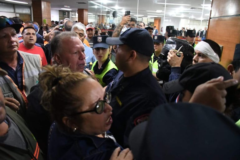 Feliciano Martínez Peña habla intensamente entre un grupo preocupado de personas en un ambiente cerrado, algunas con expresiones de protesta.