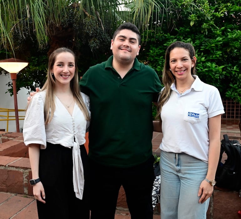 Dos mujeres y un hombre sonríen en un jardín, luciendo camisetas y pantalones. Una mujer lleva el logotipo de Plan International.