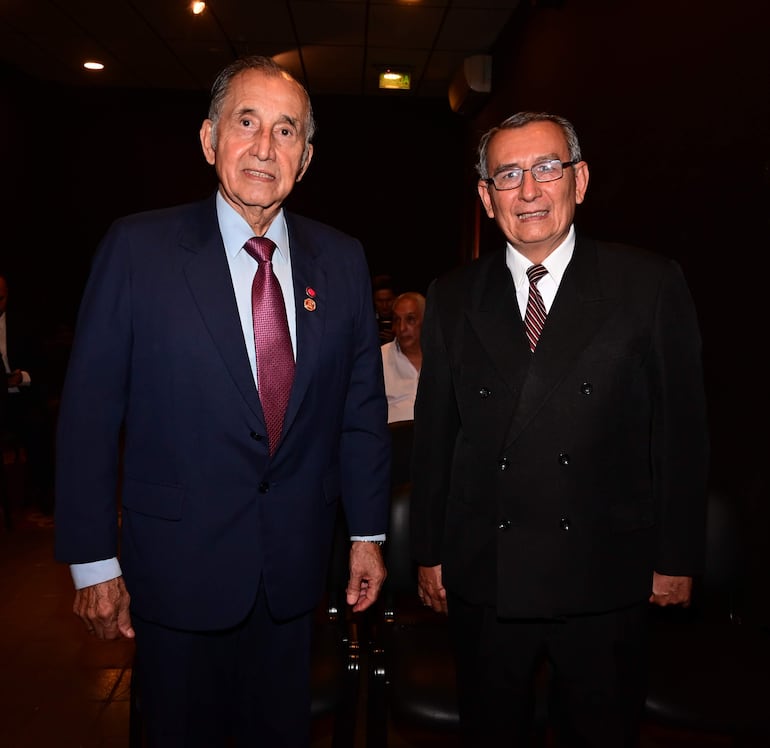 Dos hombres en trajes formales, uno con saco azul oscuro y corbata roja, el otro con traje negro y corbata a rayas, en entorno elegante.