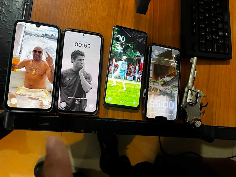 Cuatro teléfonos móviles y un revólver sobre una mesa de madera, sin personas visibles en la imagen.