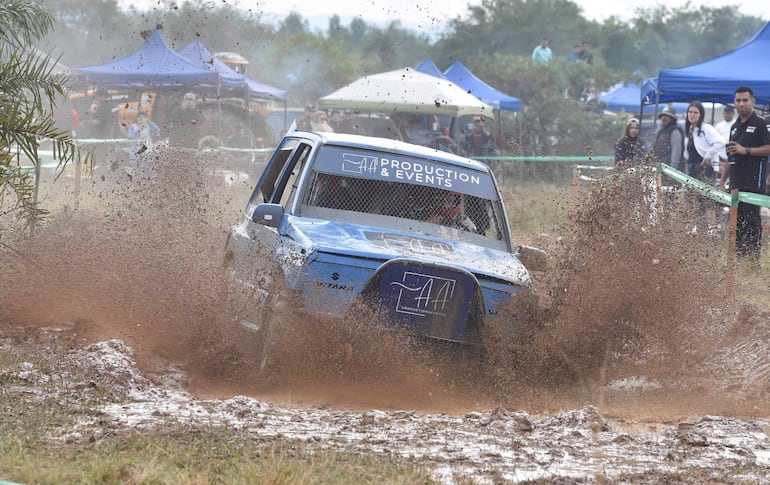 Vehículo azul avanzando a gran velocidad en terreno fangoso; ambiente de rally con carpas y público observando.